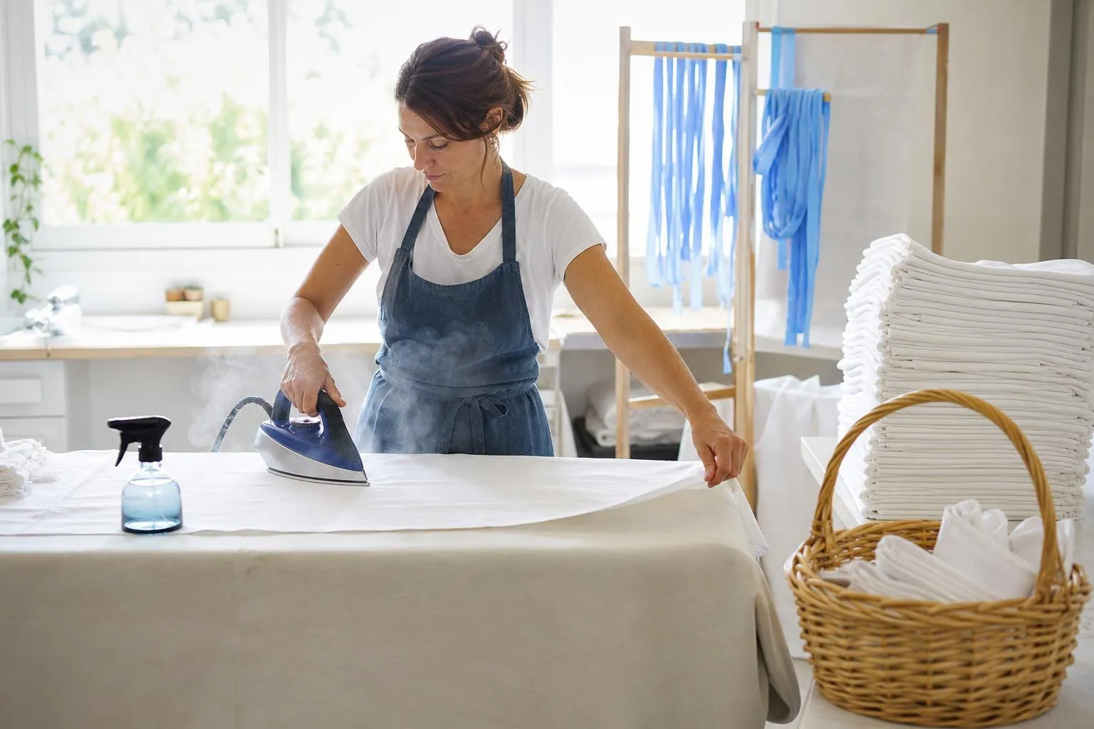 Loueuse repassant une nappe en lin blanc sur une grande table de travail, à côté d’une pile de nappes pliées et d’un panier de linge