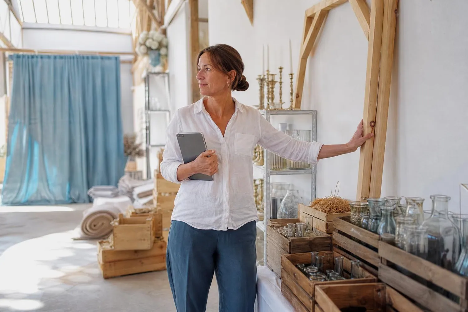 Loueuse de décoration de mariage dans son showroom, tablette à la main devant une arche en bois clair et des caisses de vases préparées pour un départ